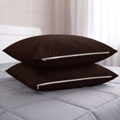 Two dark brown pillowcases stacked on a light gray quilted bedspread in a softly lit bedroom setting.