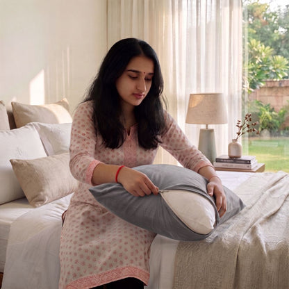 A woman in a light pink patterned kurta sits on a bed holding a gray and white zippered pillow in a bright, modern bedroom.