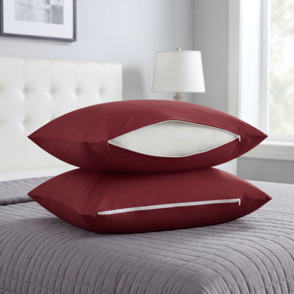 Two burgundy pillowcases stacked on a gray quilted bedspread in a bedroom setting, with a white zipper opening showing a white insert.