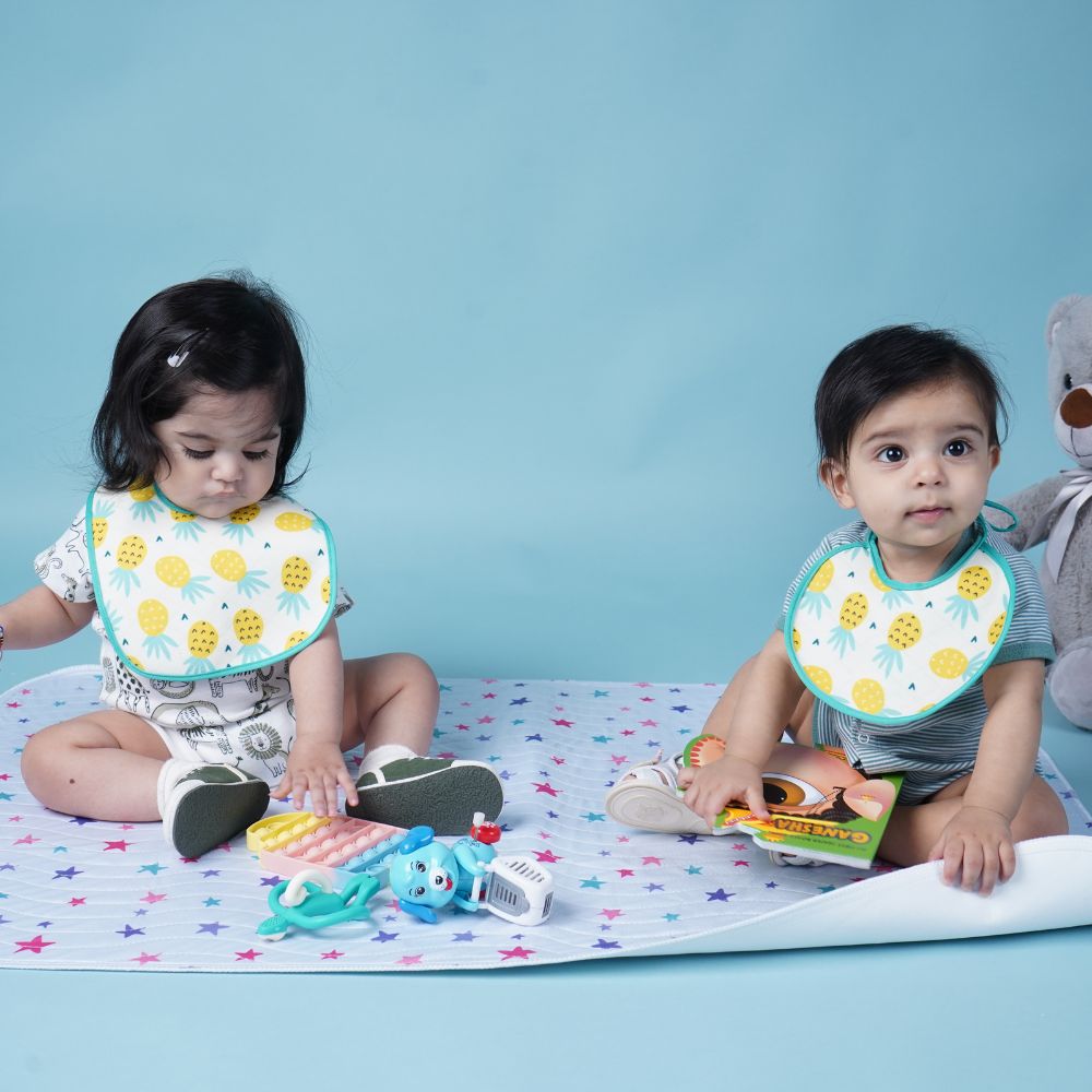 Two toddlers sitting on a pastel star blanket, wearing matching bibs with a yellow pineapple print on a white background with a blue border. The left toddler is playing with a toy, and the right toddler is holding a book, against a solid light blue background.