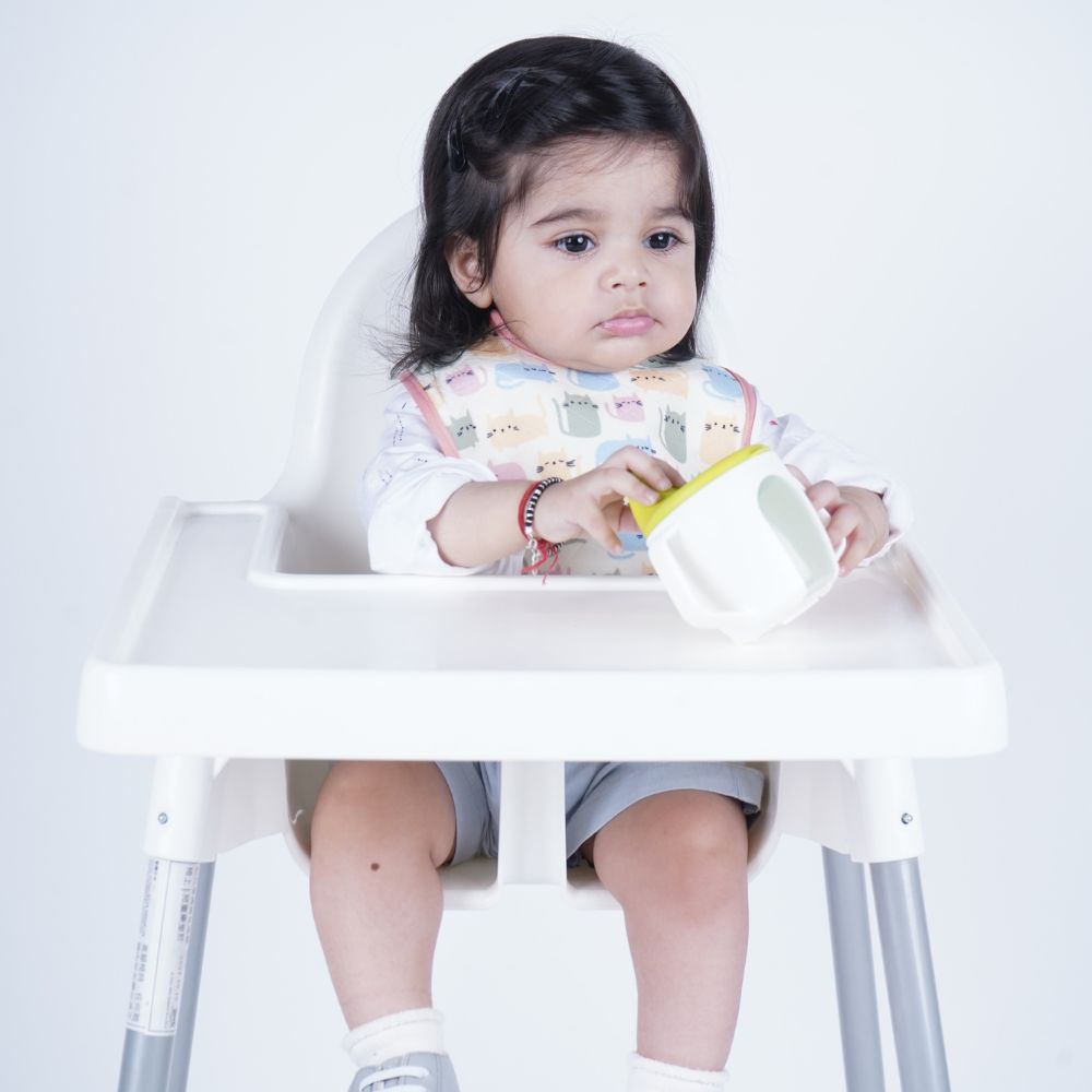 A toddler sits in a white high chair holding a white cup with a yellow lid, wearing a patterned bib and gray shorts against a plain white background.
