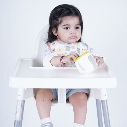 A toddler sits in a white high chair holding a white cup with a yellow lid, wearing a patterned bib and gray shorts against a plain white background.